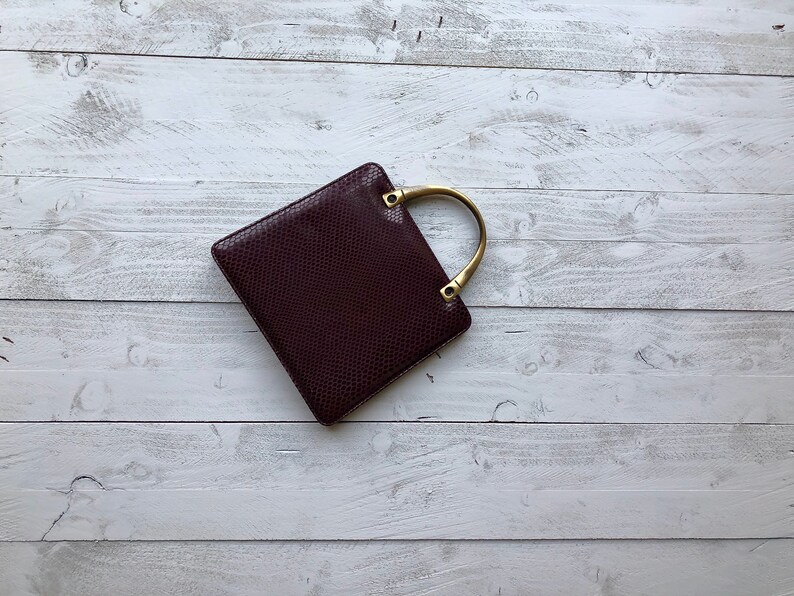May include: A small, square, burgundy handbag with a gold-toned handle. The bag has a textured, snakeskin-like pattern.