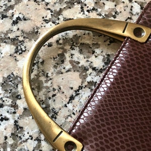 May include: A close-up of a brown leather handbag with a gold-toned handle. The handle has a decorative groove and two small rivets.