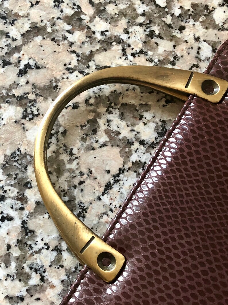 May include: A close-up of a brown leather handbag with a gold-toned handle. The handle has a decorative groove and two small rivets.