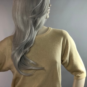 May include: A pale tan short-sleeved sweater on a mannequin. The sweater features a crew neck and a loose fit. The mannequin has long, wavy, silver hair. The background is a light grey.