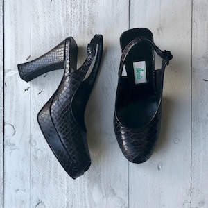 Puede incluir: Un par de sandalias de tacón de piel de serpiente negra con puntera abierta y tira de tobillo. Las sandalias tienen una suela de plataforma y un tacón alto. El nombre de la marca "Lortie" es visible en la sandalia derecha.