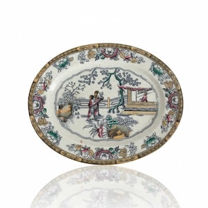 May include: An oval ceramic platter featuring a central illustration of two figures standing near a structure with a tree and rocks. The border is decorated with a repeating floral pattern in shades of pink, green, yellow, and brown on a white base.