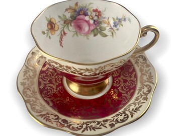 Pode incluir: Um conjunto de chá vintage com xícara e pires com um padrão floral vermelho e dourado. A xícara tem um interior branco e uma borda dourada. O pires tem uma borda ondulada.