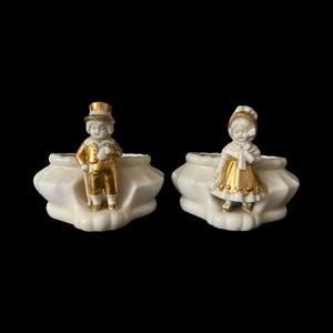 May include: A pair of white porcelain planters featuring gold-painted accents. One planter displays a figure wearing a top hat and suit, while the other features a figure in a bonnet and dress. Both pieces have a scalloped, multi-sided base design.