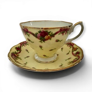 Puede incluir: Un juego de taza de té y platillo de porcelana de color amarillo pálido con un delicado patrón de rosas rosas y amarillas, hojas verdes y detalles de cintas rojas. El borde de la taza y el platillo tiene un fino acabado metálico dorado.