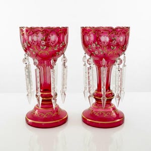 May include: A pair of ruby red antique glass goblets with gold trim and floral designs. Each goblet features dangling crystal prisms. The goblets have a wide bowl, a slender stem, and a round base. The background is white.