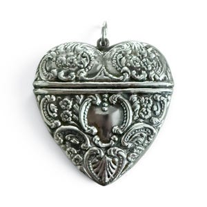 May include: A heart-shaped silver locket featuring intricate embossed floral and scrollwork patterns across the entire surface. The metal pendant has a hinged horizontal opening and a small silver loop at the top for attachment to a chain or cord.