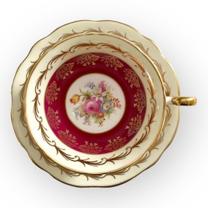 May include: Cream-colored teacup and saucer set with gold trim and a floral design. The interior of the cup is a deep red with gold filigree and a central floral bouquet. The handle and rim are accented with gold.