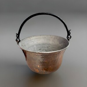 Puede incluir: Una olla de cobre con acabado martillado y un asa de metal negro. La olla tiene un fondo redondeado y una abertura ancha.