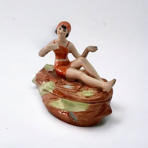 May include: A ceramic figurine of a woman in a red swimsuit and cap, reclining on a shell-shaped base. The base is coloured brown, green, and tan. The figurine has a vintage aesthetic.