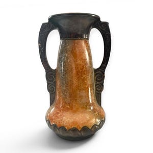May include: An antique vase with a textured, mottled orange and brown finish. The vase has a dark brown rim and base, with decorative handles on either side. The handles and the decorative elements are dark brown.
