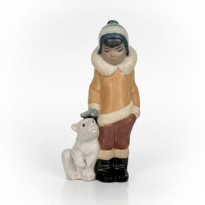 May include: A ceramic figurine of a child in a winter coat and hat, petting a polar bear cub. The child's coat is tan with white trim, and the pants are maroon. The polar bear is white, and the boots are black.