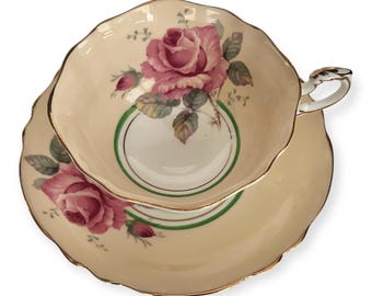 May include: A vintage teacup and saucer set with a delicate floral design. The cup and saucer are cream-colored with a green and gold trim. The floral design features pink roses with green leaves.