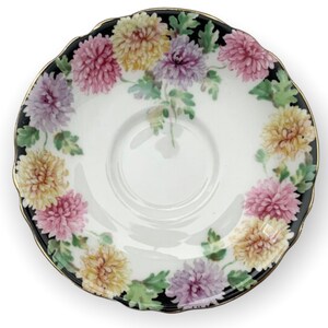 May include: A white porcelain saucer with a black rim and gold trim. The saucer is decorated with a floral pattern of pink, yellow, and purple chrysanthemums.