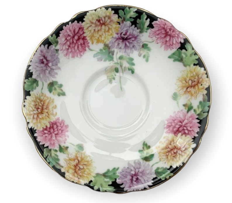 May include: A white porcelain saucer with a black rim and gold trim. The saucer is decorated with a floral pattern of pink, yellow, and purple chrysanthemums.
