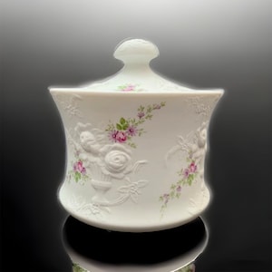 May include: White ceramic sugar bowl with a lid, featuring a raised floral design with pink roses and green leaves. The bowl has a rounded shape and a smooth, glossy finish.