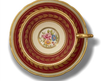 May include: A vintage teacup and saucer set with a red and gold floral design. The cup and saucer have a white background with a gold rim and a red band with a gold floral pattern.
