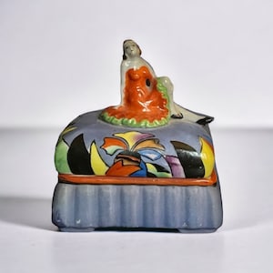 May include: A small ceramic trinket box with a blue and white base and a colourful lid. The lid features a stylised woman in a red dress sitting on a blue and white cushion.
