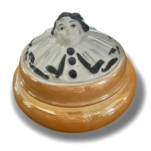 May include: A ceramic trinket box with a lid featuring a hand-painted clown with a white ruff and black accents. The box is a light brown colour with a glossy finish.