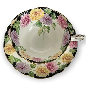 May include: A vintage teacup and saucer set with a floral design. The cup and saucer are white with a black rim and a gold trim. The floral design features pink, yellow, and white chrysanthemums on a green background.
