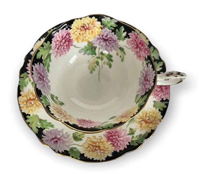 May include: A vintage teacup and saucer set with a floral design. The cup and saucer are white with a black rim and a gold trim. The floral design features pink, yellow, and white chrysanthemums on a green background.