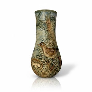 May include: A ceramic vase featuring a relief design of a brown bird standing near a nest with three eggs. The surface displays a muted colour palette of blue, green, and brown tones with a textured finish depicting tall grasses and foliage.