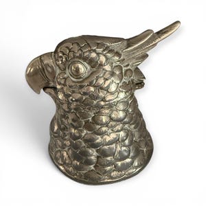 May include: A metallic, silver-coloured parrot head figurine featuring detailed, embossed feather textures across the entire surface. The bird has a prominent curved beak, a defined eye, and a crest of feathers extending from the top of its head.