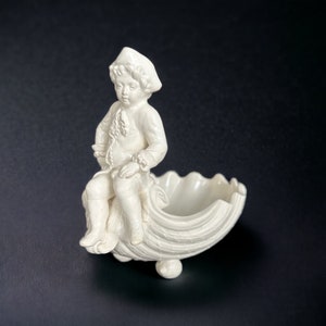 May include: A white ceramic figurine of a boy sitting on a shell-shaped dish. The boy is wearing a hat and a jacket. The dish has a scalloped edge and three feet.