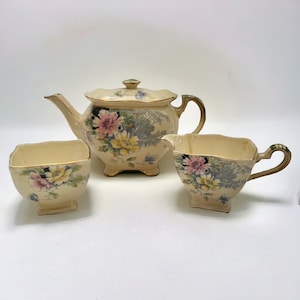 May include: A vintage tea set with a cream-colored base and floral pattern. The set includes a teapot, creamer, and sugar bowl.
