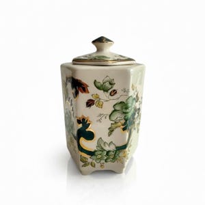 May include: Cream-coloured ceramic jar with a lid, featuring floral designs in green, gold, and brown. The jar has a hexagonal shape and a gold trim. The lid is topped with a dark green finial.