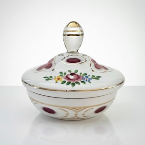 Może przedstawiać: Biała ceramiczna miska z pokrywką, złotym uchwytem i wykończeniem. Pokrywka ma kwiatowy wzór z centralną różą i bordowymi akcentami. Miska ma dekoracyjny wzór z bordowych kropek i złotych linii.