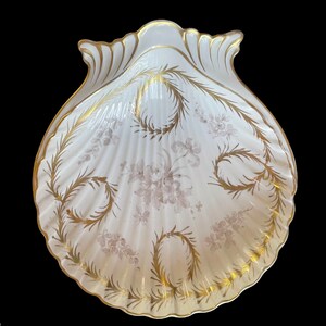 Könnte beinhalten: Eine weiße Porzellanschale in Muschelform mit goldenen und grauen floralen Designs. Die Schale ist perfekt zum Servieren kleiner Snacks oder Desserts.