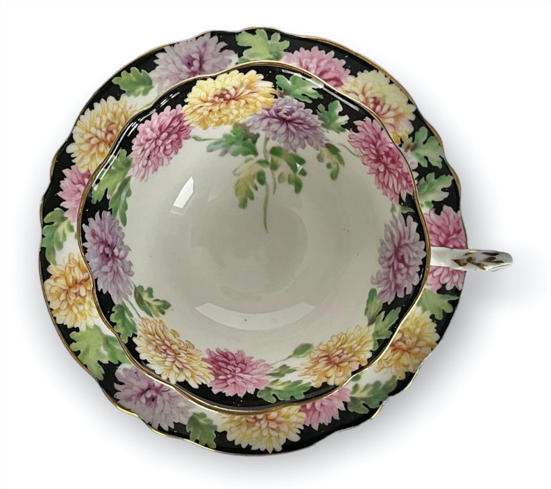 May include: A vintage teacup and saucer set with a black background and a floral design featuring pink, yellow, and white chrysanthemums.