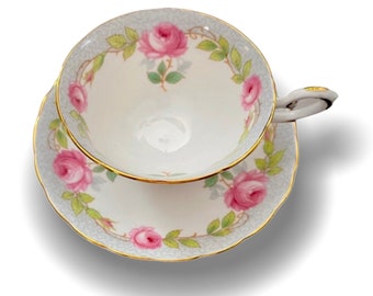 May include: A white teacup and saucer set with a delicate floral pattern. The cup and saucer feature a gray background with a gold trim and pink roses with green leaves.
