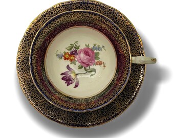 May include: A vintage teacup and saucer set featuring a floral design. The cup and saucer are decorated with a black and gold pattern. The floral design includes pink, yellow, and purple flowers.