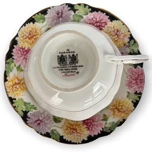 May include: A white teacup and saucer with a black and white floral pattern. The saucer has a gold rim. The teacup has a white interior and a black and white floral pattern on the exterior. The teacup has a handle and the saucer has a scalloped edge. The teacup has a mark on the bottom that reads "Paragon by Appointment to HM Queen Mary Fine Bone China England Regd.  Aucuna."