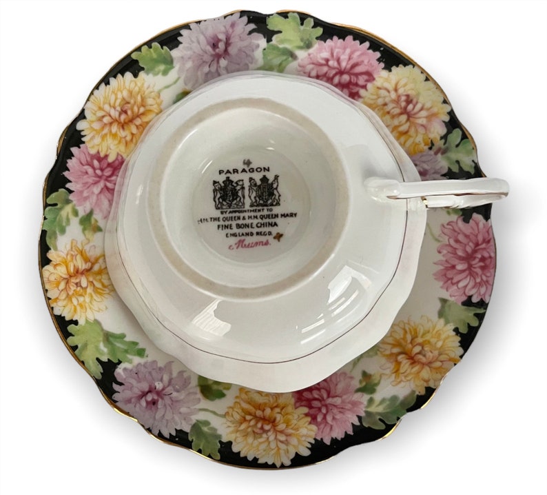 May include: A white teacup and saucer with a black and white floral pattern. The saucer has a gold rim. The teacup has a white interior and a black and white floral pattern on the exterior. The teacup has a handle and the saucer has a scalloped edge. The teacup has a mark on the bottom that reads "Paragon by Appointment to HM Queen Mary Fine Bone China England Regd.  Aucuna."