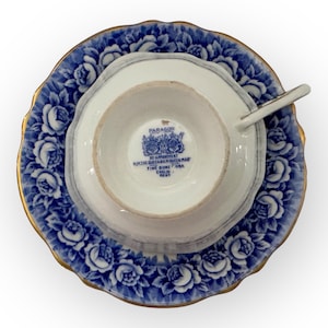 May include: A blue and white teacup and saucer set with a gold rim. The saucer features a floral pattern with blue roses. The teacup has a gold rim and the words "Paragon by Appointment to HM The Queen Mother Fine Bone China England Regd" on the bottom.
