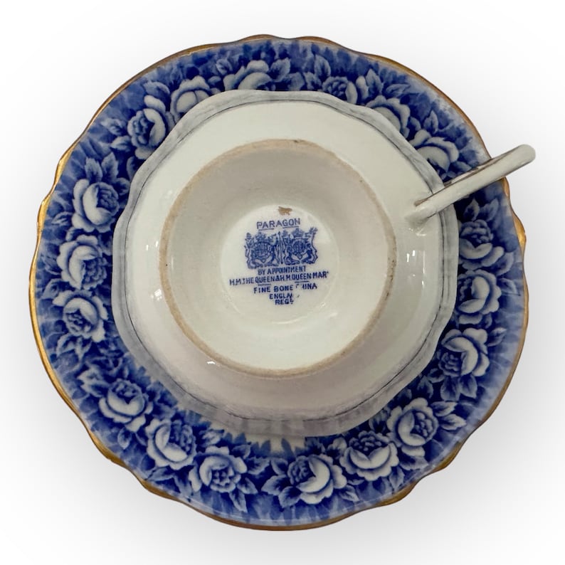 May include: A blue and white teacup and saucer set with a gold rim. The saucer features a floral pattern with blue roses. The teacup has a gold rim and the words "Paragon by Appointment to HM The Queen Mother Fine Bone China England Regd" on the bottom.