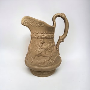 Puede incluir: Una jarra de cerámica marrón con asa y un diseño en relieve de una persona montando a caballo. La jarra tiene un diseño detallado y un aspecto rústico.