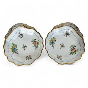 May include: Two white, scalloped-edge porcelain dishes with floral designs. Each dish features a blue border, gold trim, and hand-painted flowers in shades of pink, yellow, blue, and purple. The dishes have a decorative, ornate design.