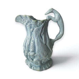May include: A light blue ceramic jug featuring a relief design of rabbits and foliage. The handle is shaped like a greyhound dog. The jug has a scalloped rim and a textured surface with a glossy finish, showcasing intricate sculptural details.