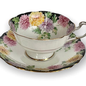 May include: A white teacup and saucer with a black rim and a floral pattern of pink, yellow, and purple chrysanthemums.