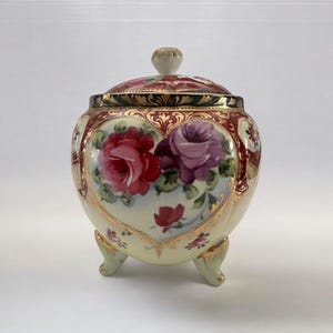 May include: A heart-shaped porcelain jar with a lid, decorated with hand-painted roses in shades of pink, purple, and red. The jar has gold accents and sits on three small feet.
