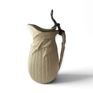 May include: A cream-coloured ceramic pitcher featuring a textured surface that mimics the appearance of corn kernels and husks. The pitcher includes a dark metal hinged lid and a matching handle shaped like a stalk, providing a functional design.