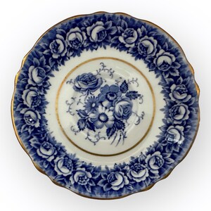 May include: A white porcelain saucer with a blue and gold floral design. The saucer has a scalloped edge and features a delicate floral pattern with roses and leaves.