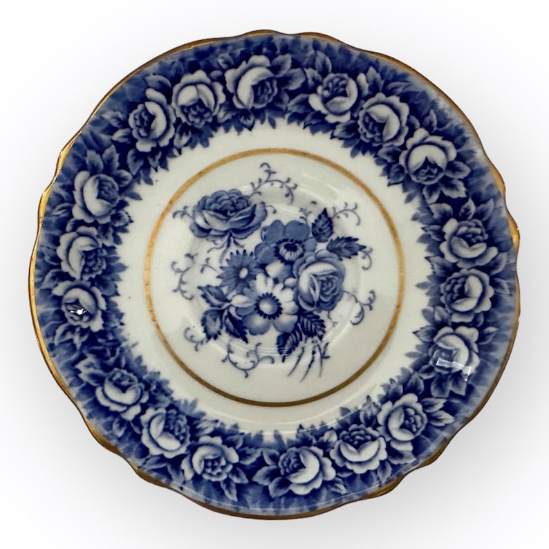 May include: A white porcelain saucer with a blue and gold floral design. The saucer has a scalloped edge and features a delicate floral pattern with roses and leaves.