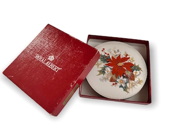 May include: A Royal Albert Poinsettia pattern plate in a red box. The plate features a central poinsettia flower design with white flowers, green leaves, and gold trim. The red box has the Royal Albert logo.