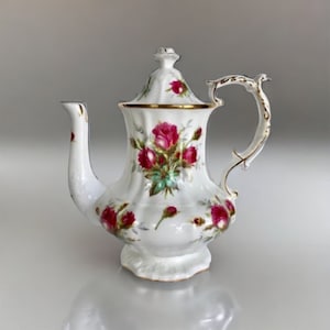 Może przedstawiać: Biały porcelanowy czajnik z kwiatowym wzorem z różowymi i czerwonymi różami. Czajnik ma złotą obwódkę wokół pokrywki i uchwytu.