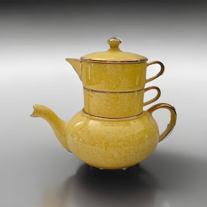 Könnte beinhalten: Eine gelbe Teekanne aus Keramik mit goldener Verzierung. Die Teekanne hat einen runden Korpus und einen Ausguss an der Seite. Die Teekanne hat einen Deckel mit einem Knopf oben. Die Teekanne ist auf zwei kleineren Tassen gestapelt, ebenfalls mit goldener Verzierung.